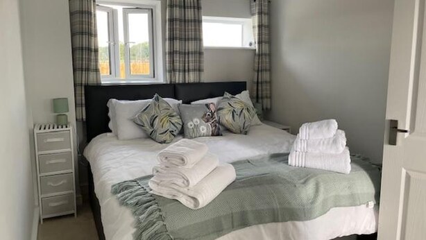 A neatly made bed with pillows and a blanket, a bedside table with a lamp, and a window with curtains. - Barn Retreat/ Hottub/ Bbq/ Petsok/wi-fi (Evesham)