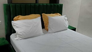 Un letto con biancheria bianca e due cuscini, uno bianco e uno giallo, in una stanza con una testiera imbottita verde e un grande specchio sopra il letto.
