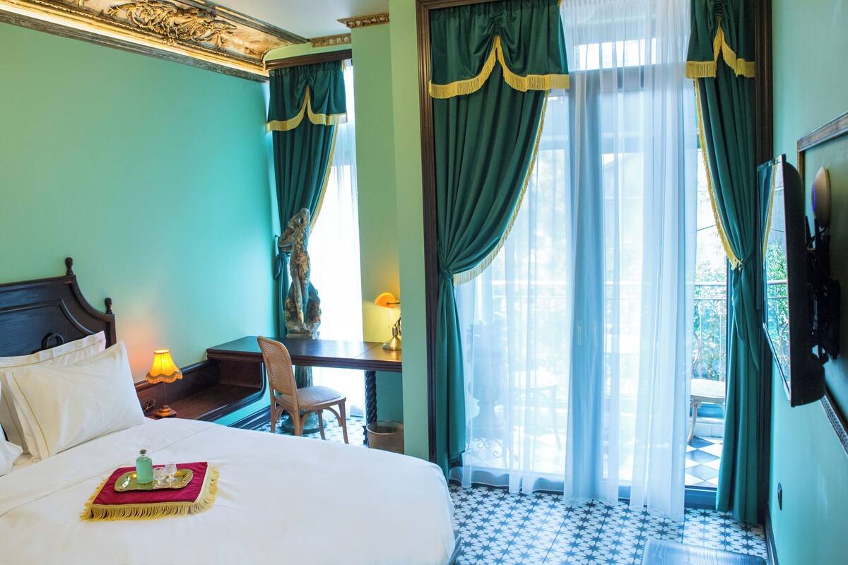 A hotel room with a large bed, a desk, and a chair. There is a window with curtains and a view of the outside.