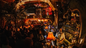 A crowded bar with a bartender, shelves of bottles, and ornate decor. - Juan Rodriguez Hacienda (Athens)