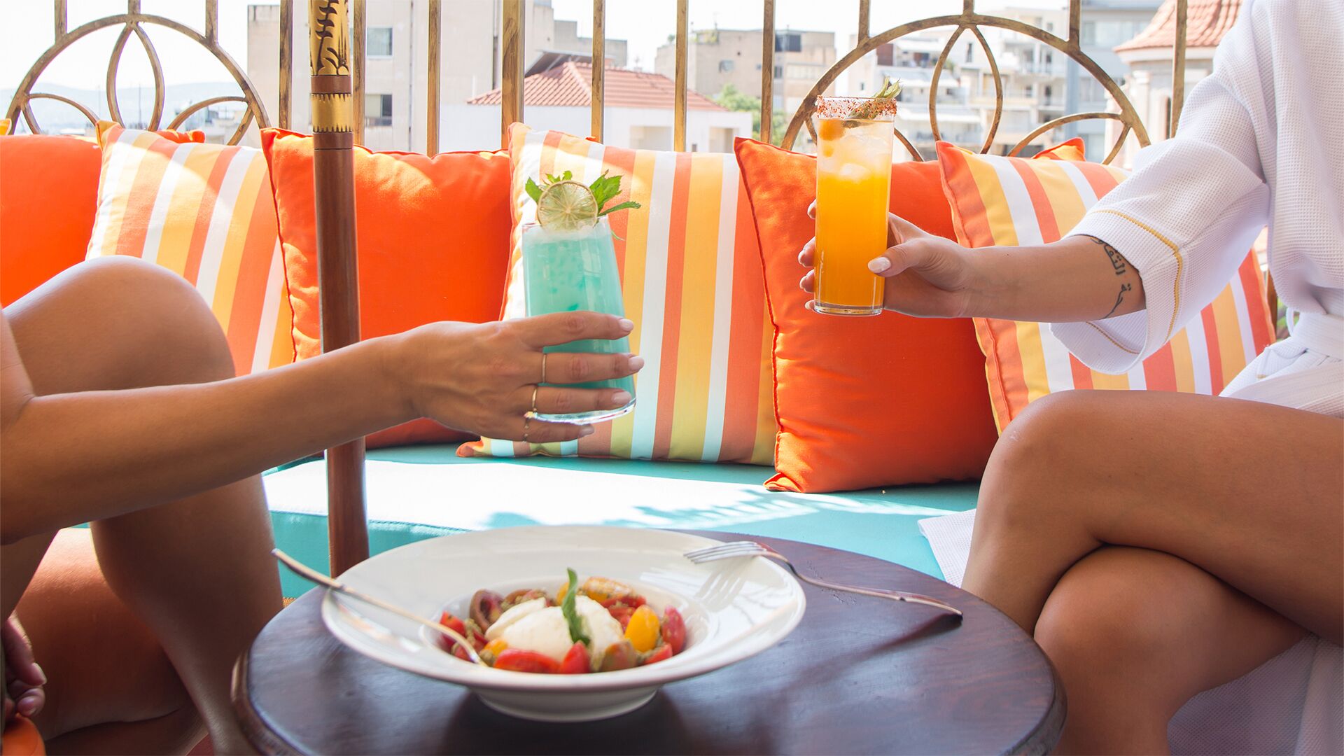 A person holding a glass of orange juice, another holding a blue drink, and a plate of food on a table.