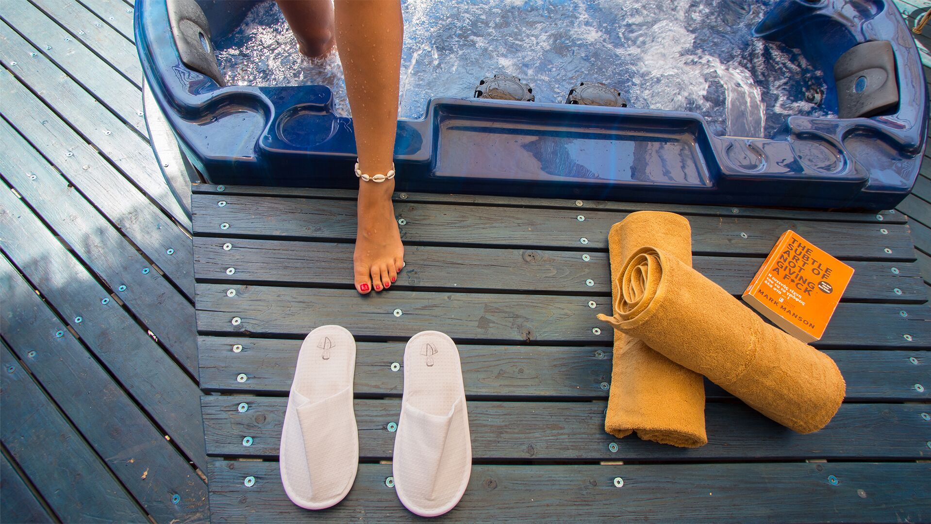 A wooden deck with a hot tub, a pair of white flip-flops, and a rolled-up towel with a book titled "The Subtle Art of Not Giving a F*ck" by Mark Manson.