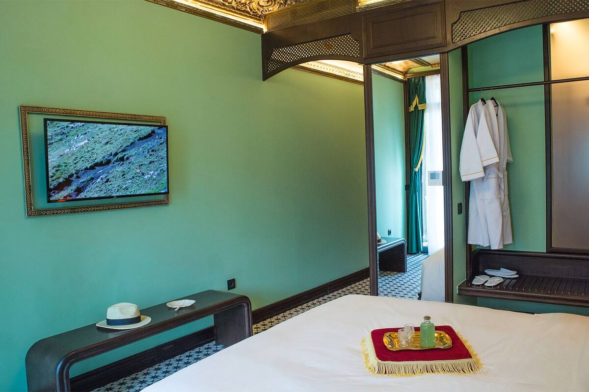 A hotel room with a bed, a bedside table with a hat and a bowl, a television on the wall, and a mirror reflecting a robe hanging on the wall.