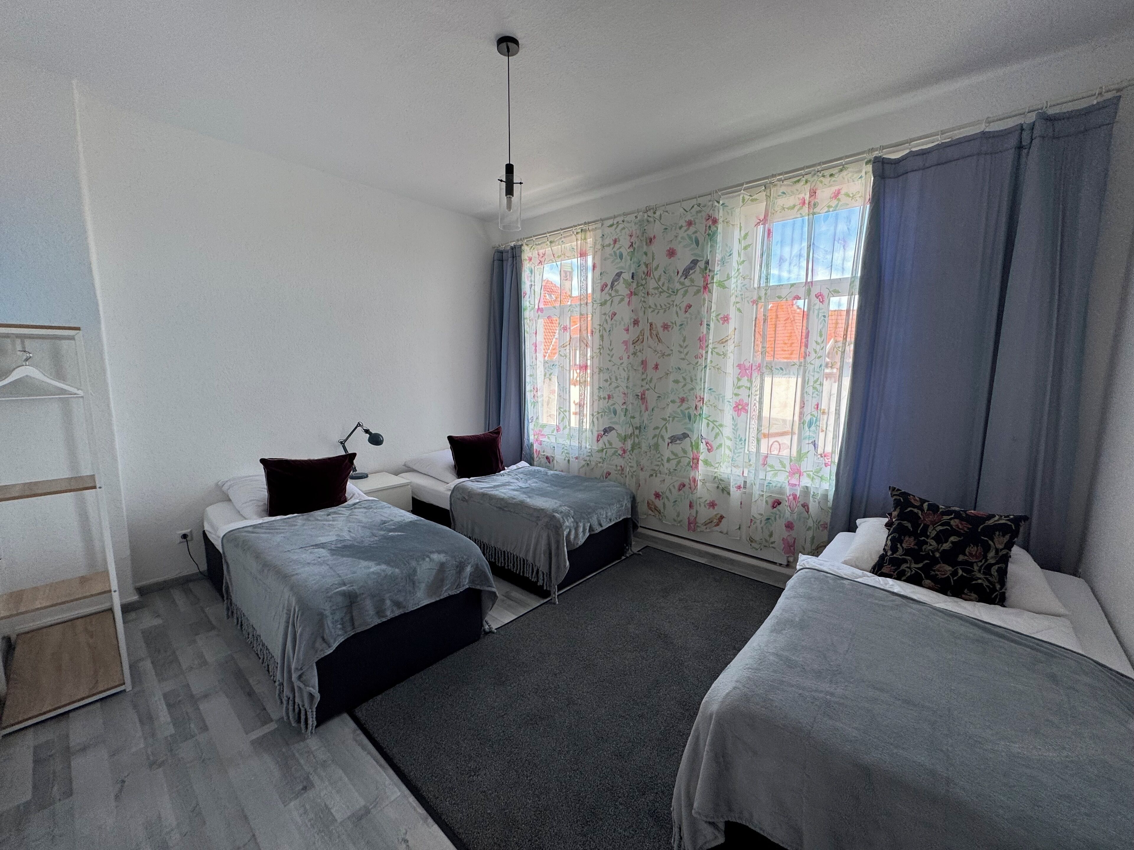 A room with two single beds, a desk with a chair, a hanging coat rack, and a window with floral curtains.