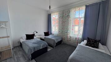 A room with two single beds, a desk with a chair, a hanging coat rack, and a window with floral curtains.