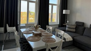 Ein modernes Wohnzimmer mit einem Esstisch, weißen Stühlen, einem grauen Sofa und Blick auf eine von Bäumen gesäumte Straße durch große Fenster.