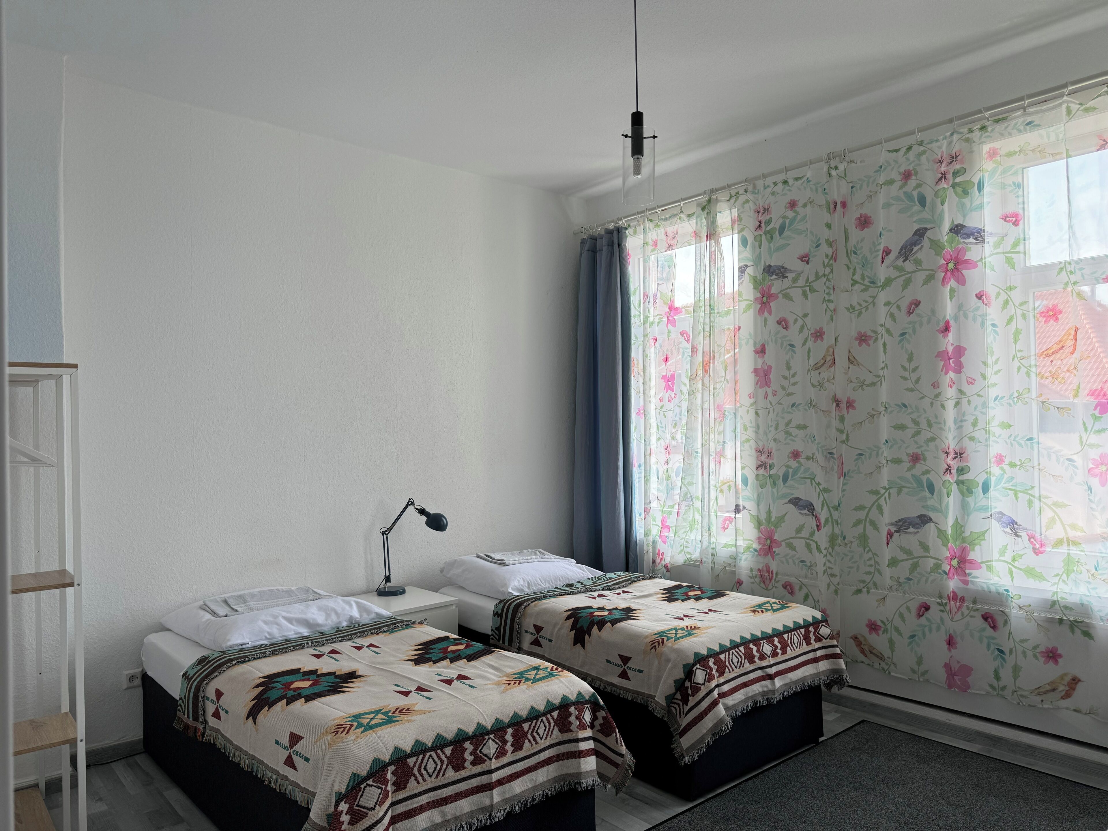 Een slaapkamer met twee bedden, een bureau, een stoel, een lamp en een raam met bloemen gordijnen.
