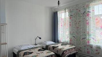 Een slaapkamer met twee bedden, een bureau, een stoel, een lamp en een raam met bloemen gordijnen.