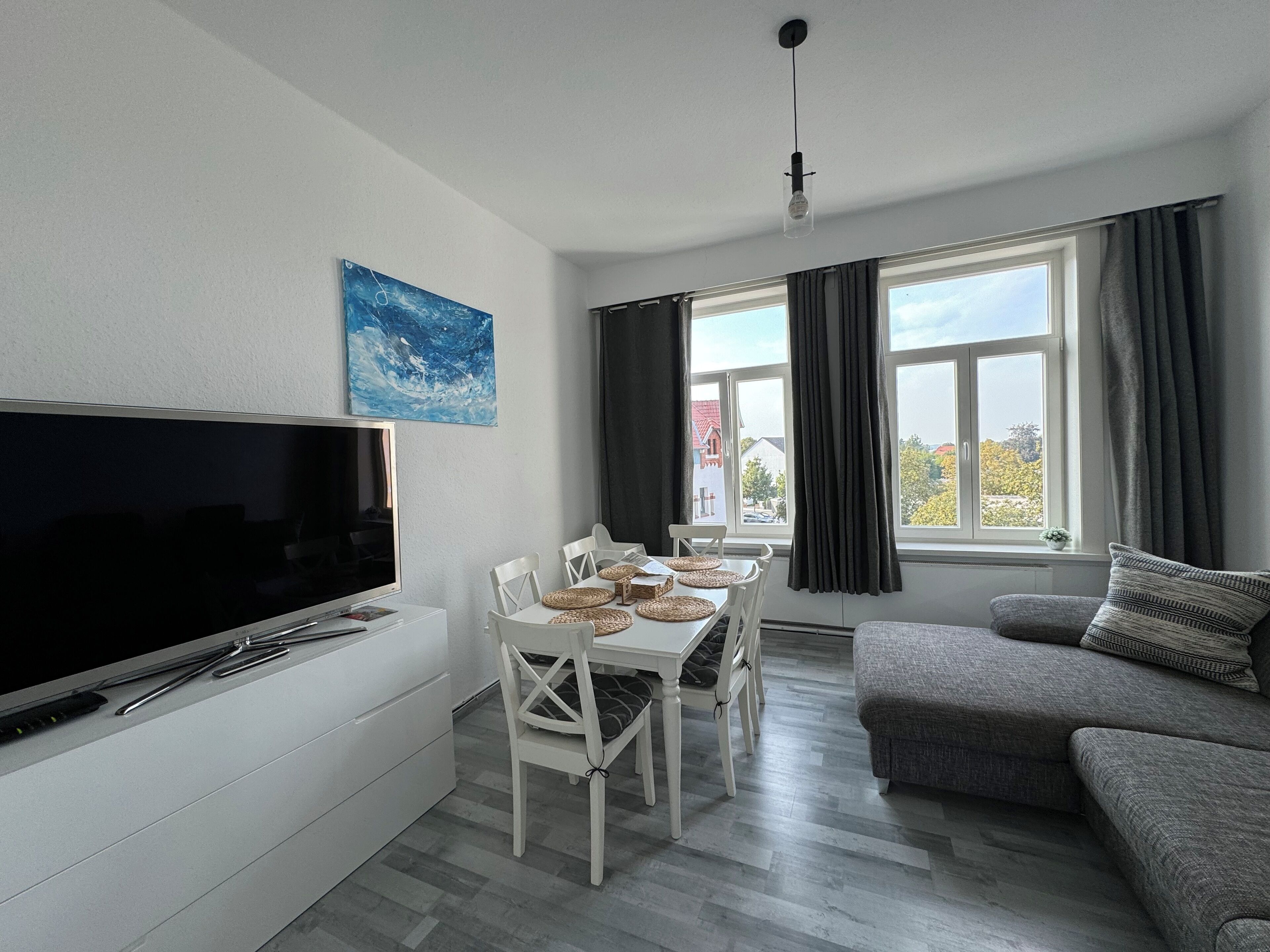 A modern living room with a flat-screen TV, a dining table with chairs, a grey sofa, and a large window with curtains.