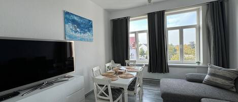 A modern living room with a flat-screen TV, a dining table with chairs, a grey sofa, and a large window with curtains.