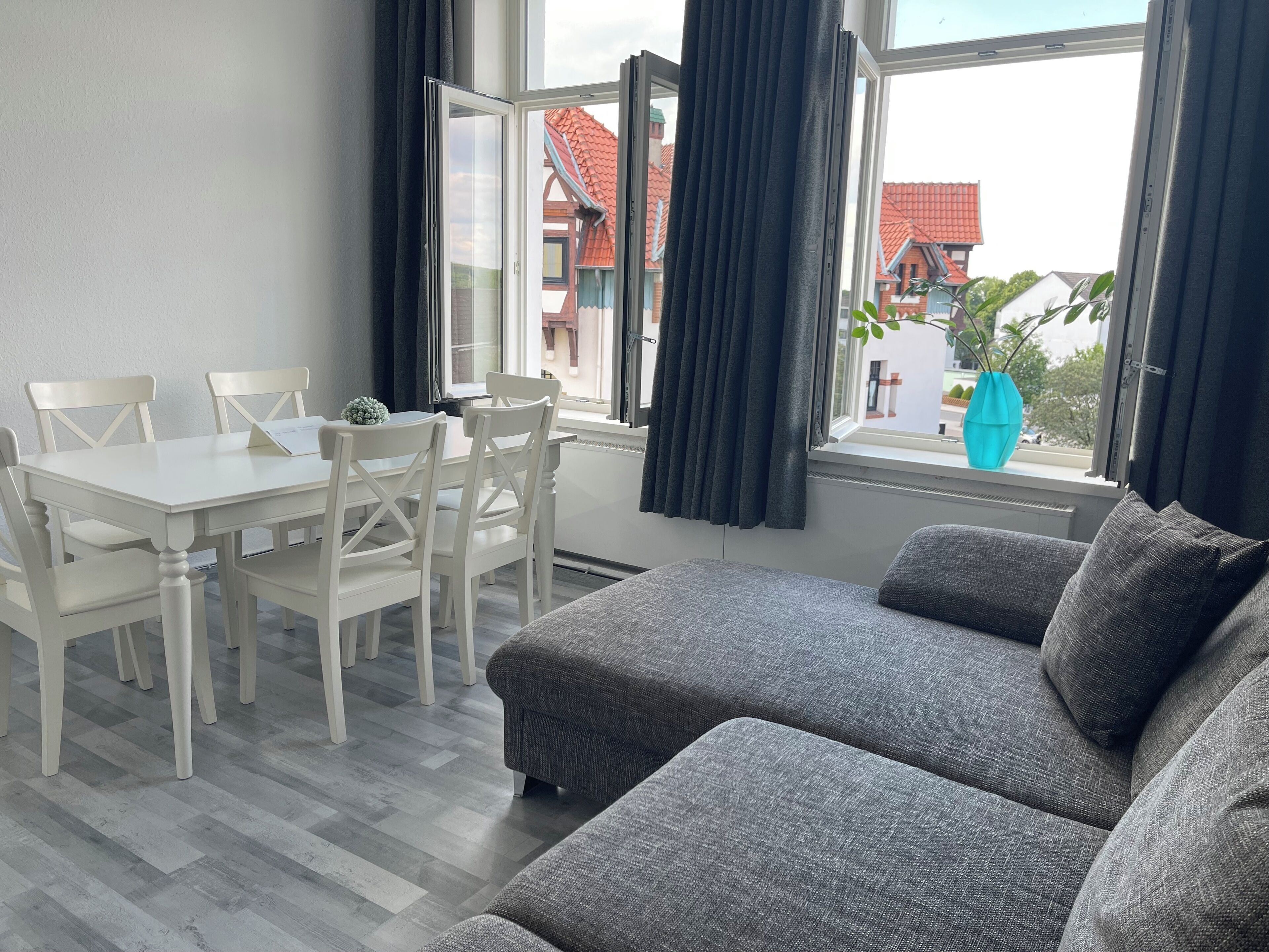 Un soggiorno moderno con un divano grigio, un tavolo da pranzo bianco e sedie. Si vede un'ampia finestra con tende, da cui si gode una vista di edifici.