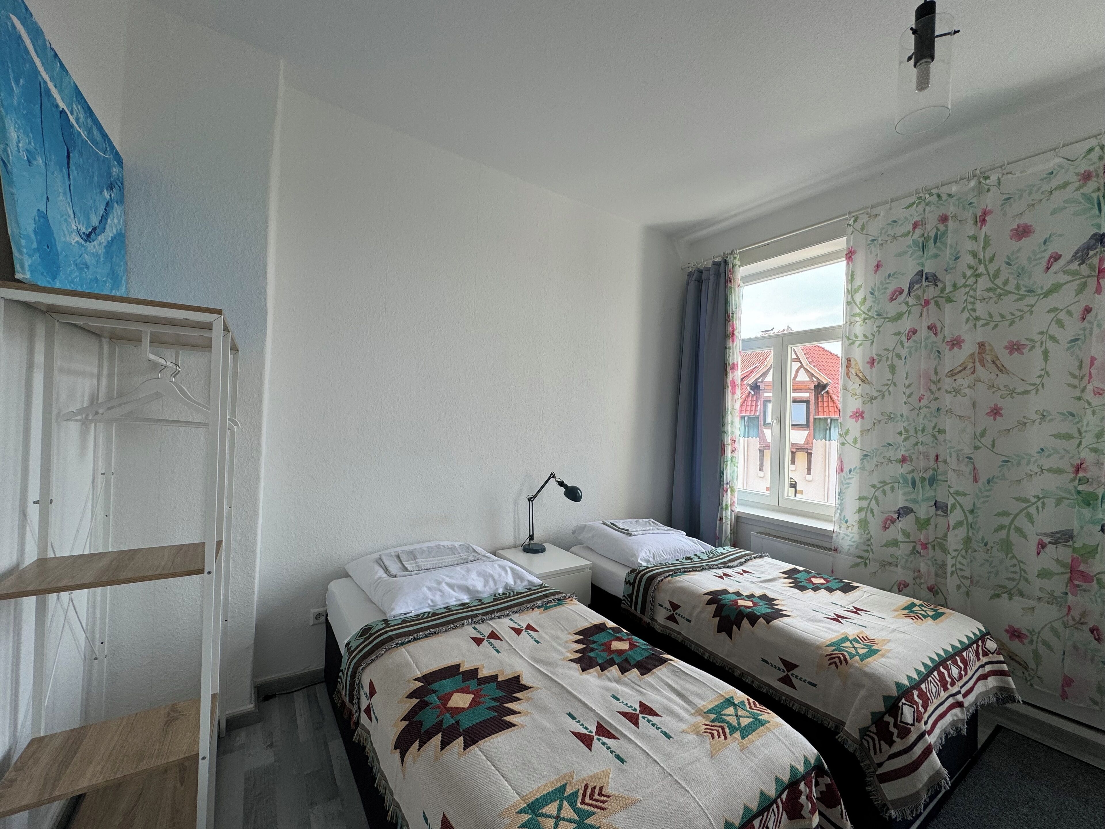 Une chambre avec deux lits, un luminaire suspendu, une fenêtre ornée de rideaux à motifs floraux et une armoire blanche.
