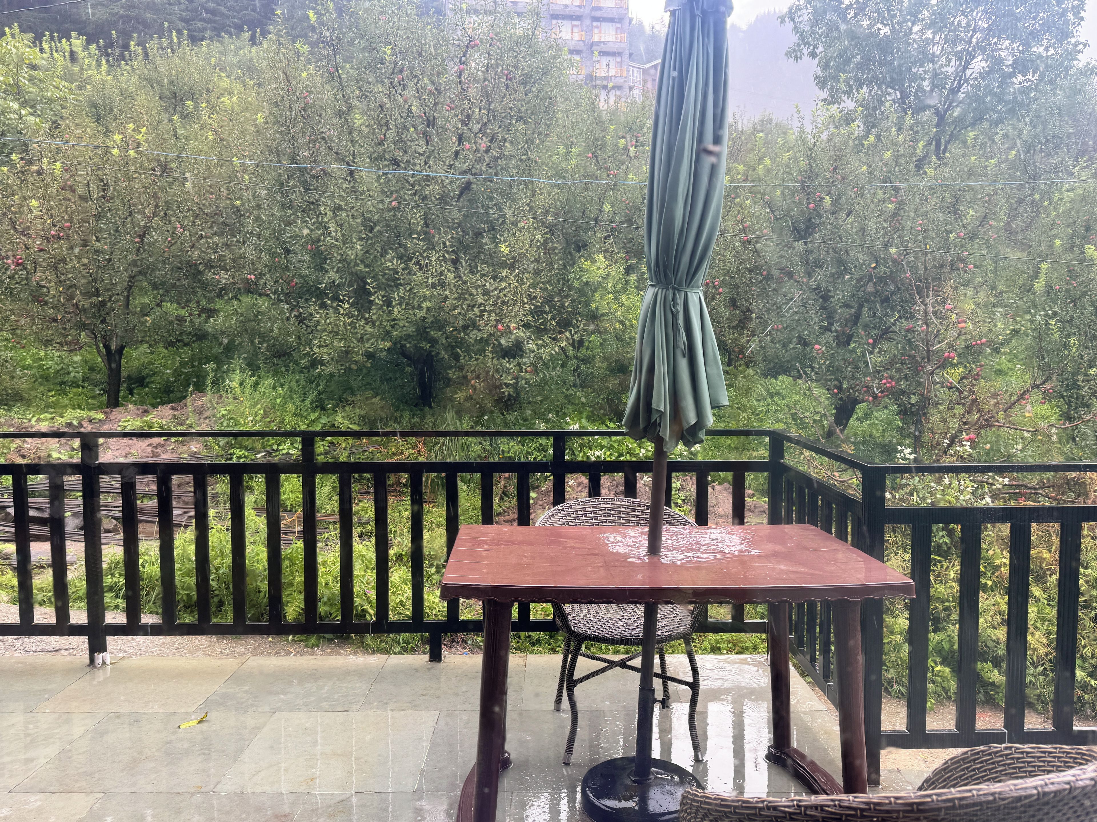 A covered outdoor seating area with a table and chairs, surrounded by a black railing, overlooking a garden with trees and a building in the background.