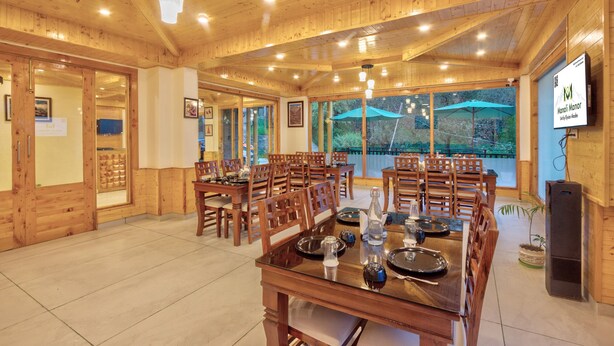 A wooden dining area with tables and chairs, a large window with an umbrella, and a sign for "Manali Manor." - Manali Manor (Manali)