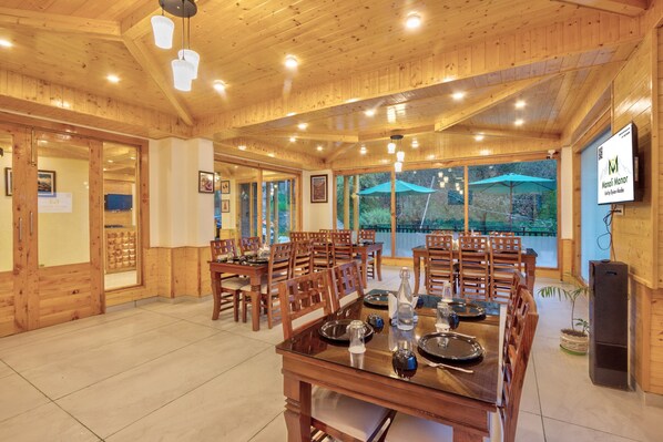 A wooden dining area with tables and chairs, a large window with an umbrella, and a sign for "Manali Manor." - Manali Manor (Manali)