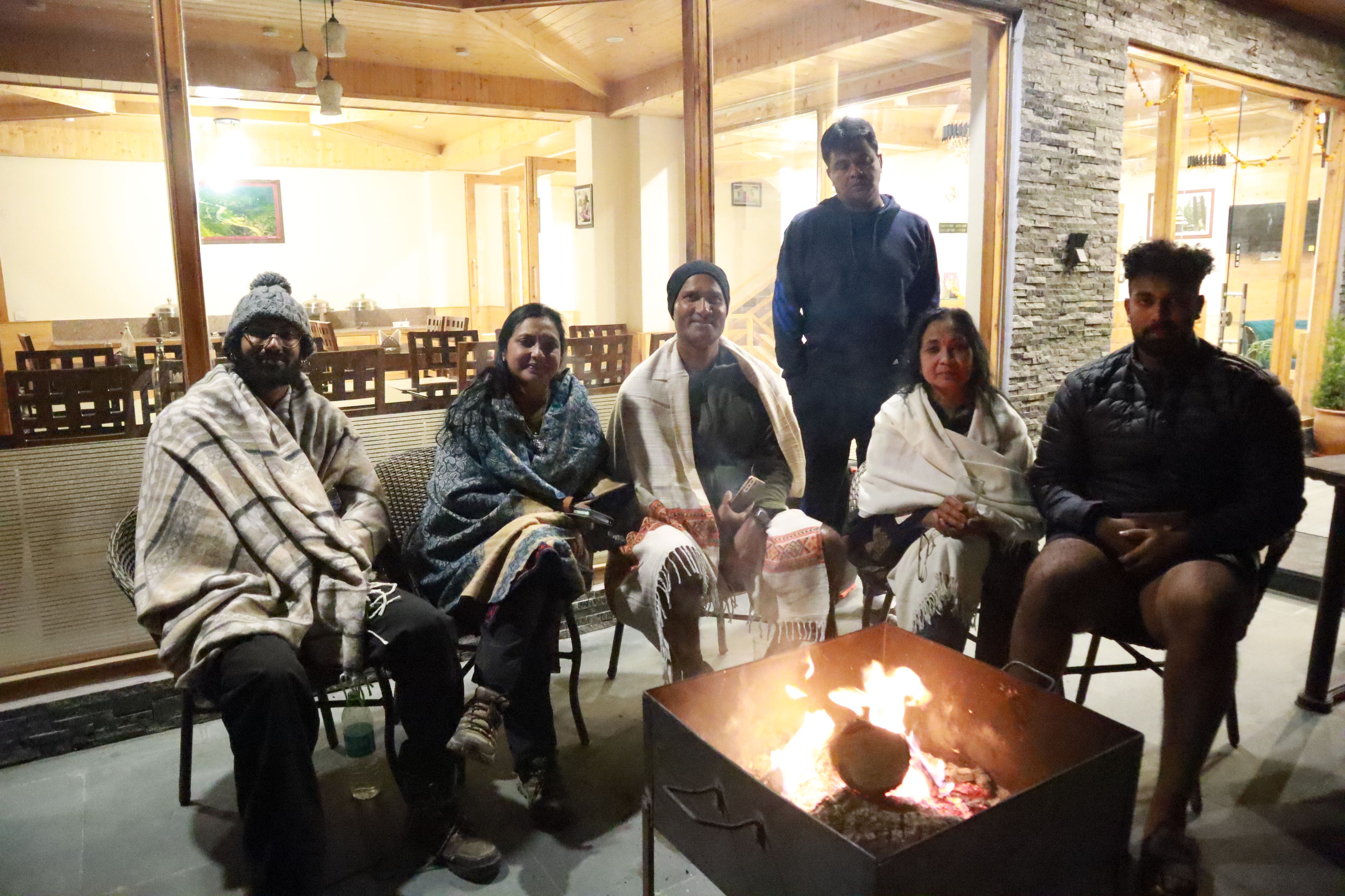 Un grupo de personas sentadas alrededor de un brasero en un espacio interior con luz cálida, muebles de madera y paredes de piedra.