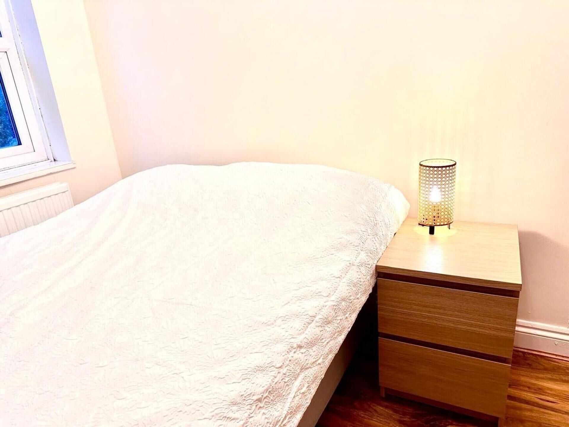 Un lit bien fait, recouvert d’une couette blanche, une table de chevet avec une lampe et une fenêtre avec des stores.