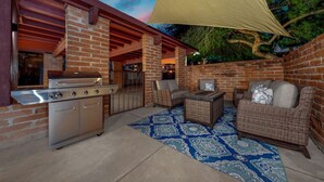 An outdoor patio with a stainless steel grill, wicker furniture, and a blue patterned rug.