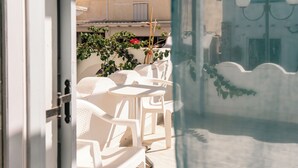Een open deur die leidt naar een terras met witte kunststof stoelen en een tafel, omgeven door potplanten en een gevel.