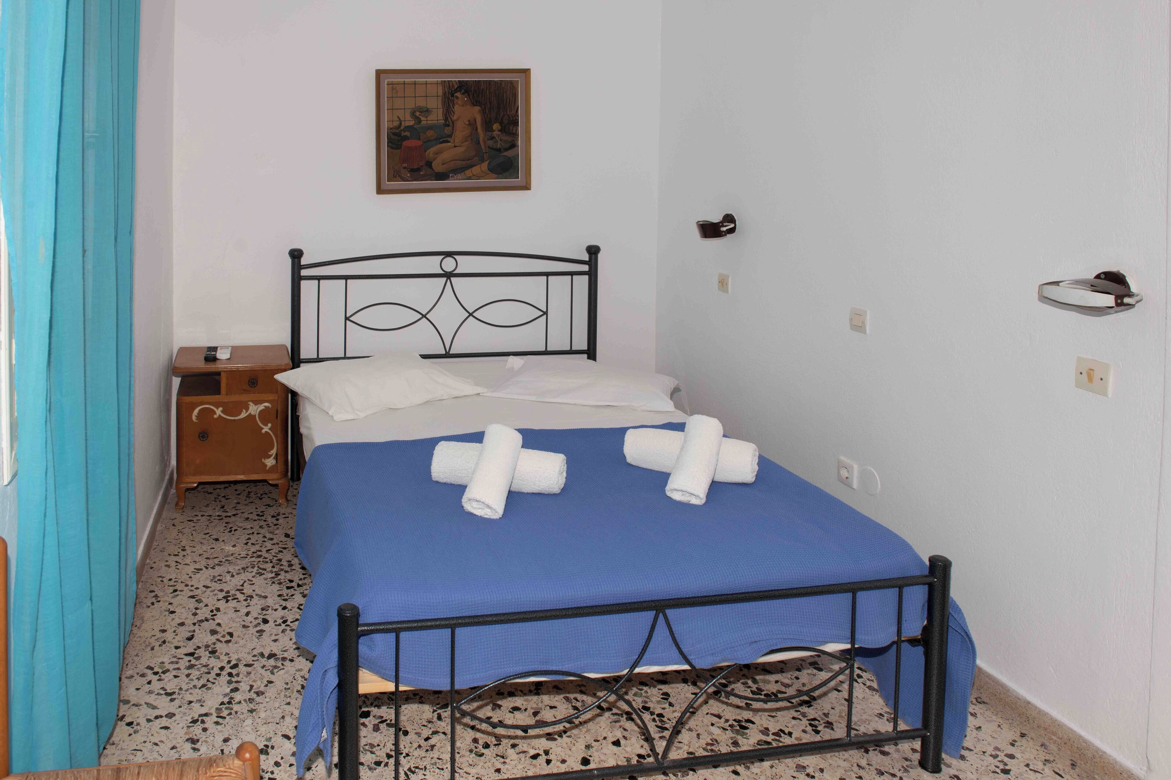 A single bed with a blue bedspread and white pillows, a wooden nightstand with a small drawer, a painting on the wall, and a blue curtain on the left.