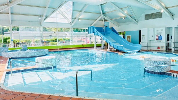 An indoor swimming pool with a blue slide, tiled steps, and a clear view of the outside. - Caravan-blackpool-familyfriendly-activities (Fleetwood)