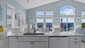 A kitchen with a large island, white cabinetry, and a view of the ocean through large windows. - Hana Kai #173 - Oceanfront Home in the Heart of Surf City! (Surf City)
