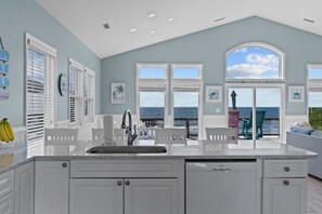A kitchen with a large island, white cabinetry, and a view of the ocean through large windows. - Hana Kai #173 - Oceanfront Home in the Heart of Surf City! (Surf City)