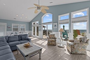 A spacious living area with a large sofa, two armchairs, a coffee table, and a ceiling fan. There are multiple windows offering a view of the ocean. - Hana Kai #173 - Oceanfront Home in the Heart of Surf City! (Surf City)