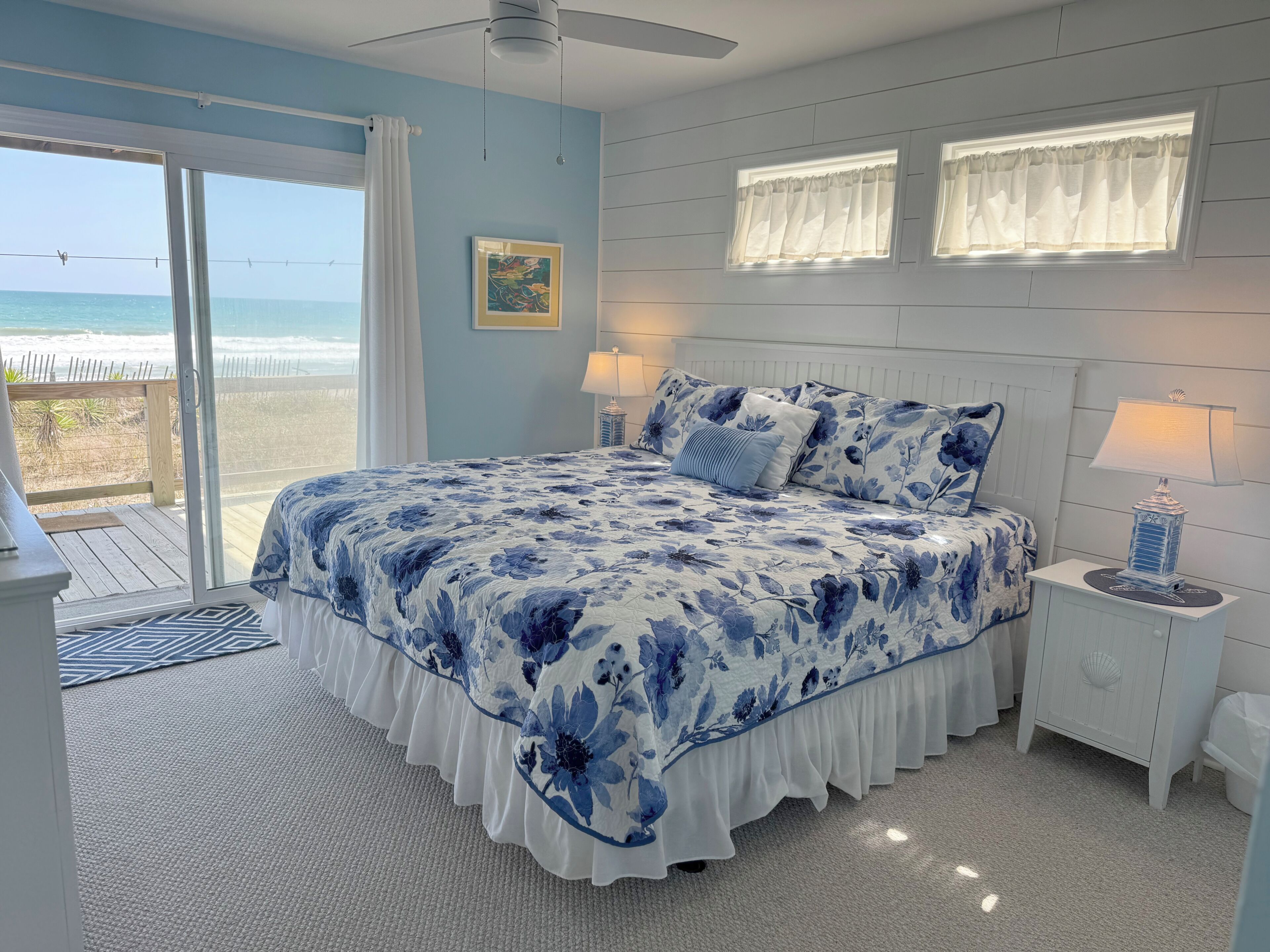 A bedroom with a large bed, two bedside lamps, a window with curtains, and a view of the beach.