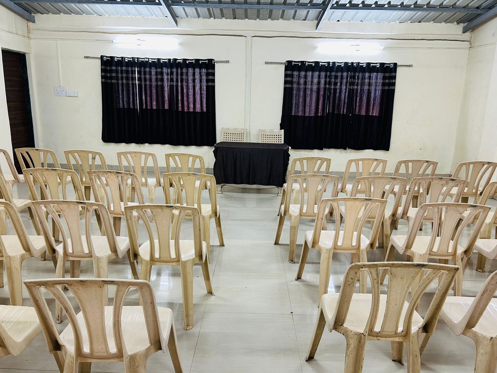 Ein Konferenzraum mit Reihen beige-farbener Kunststoffstühle, einem schwarzen Tisch im Vordergrund und schwarzen Vorhängen mit violettem Muster.