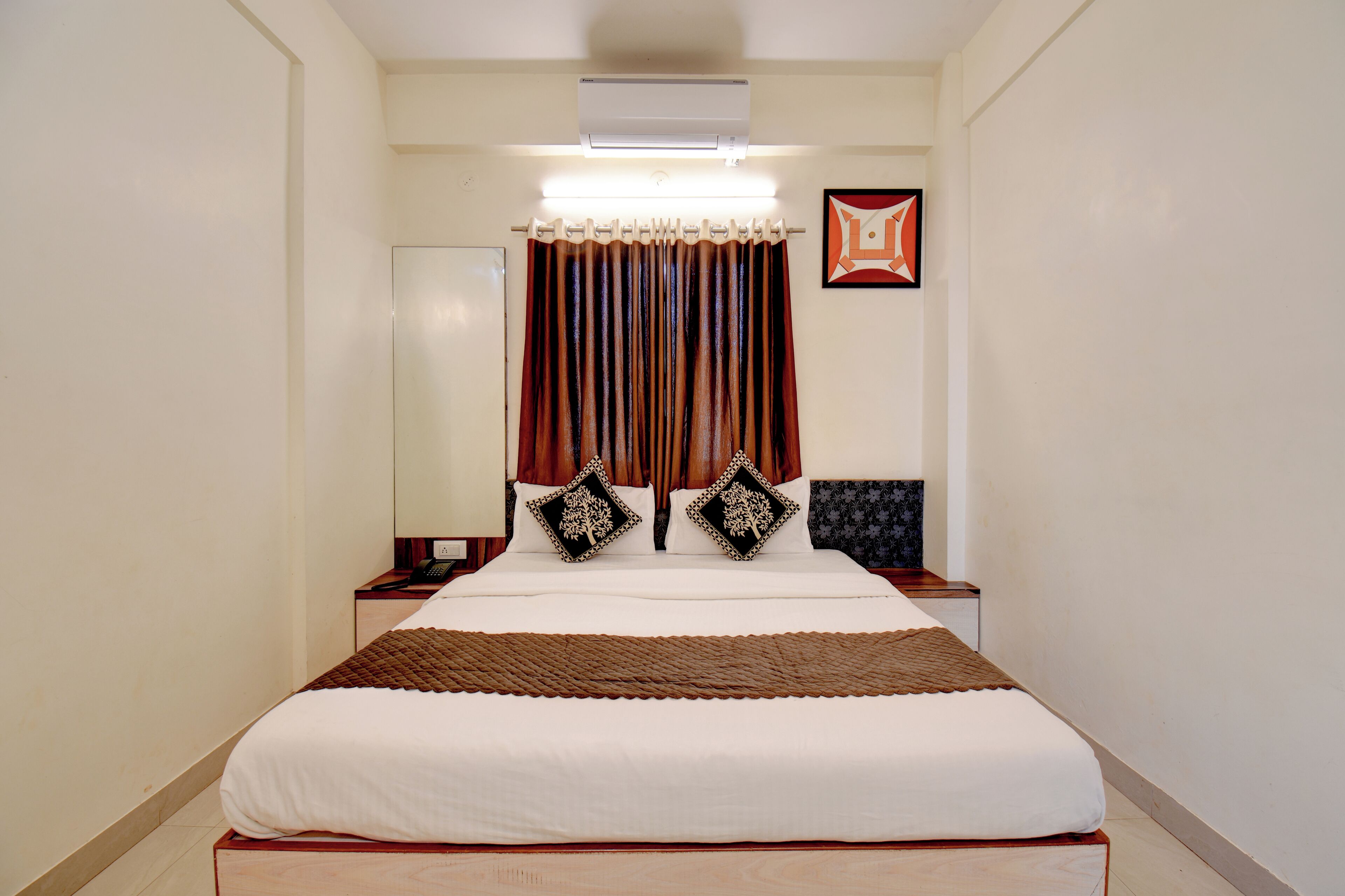 A hotel room with a large bed, a bedside table, a nightstand, a wall-mounted air conditioner, and a framed picture on the wall.