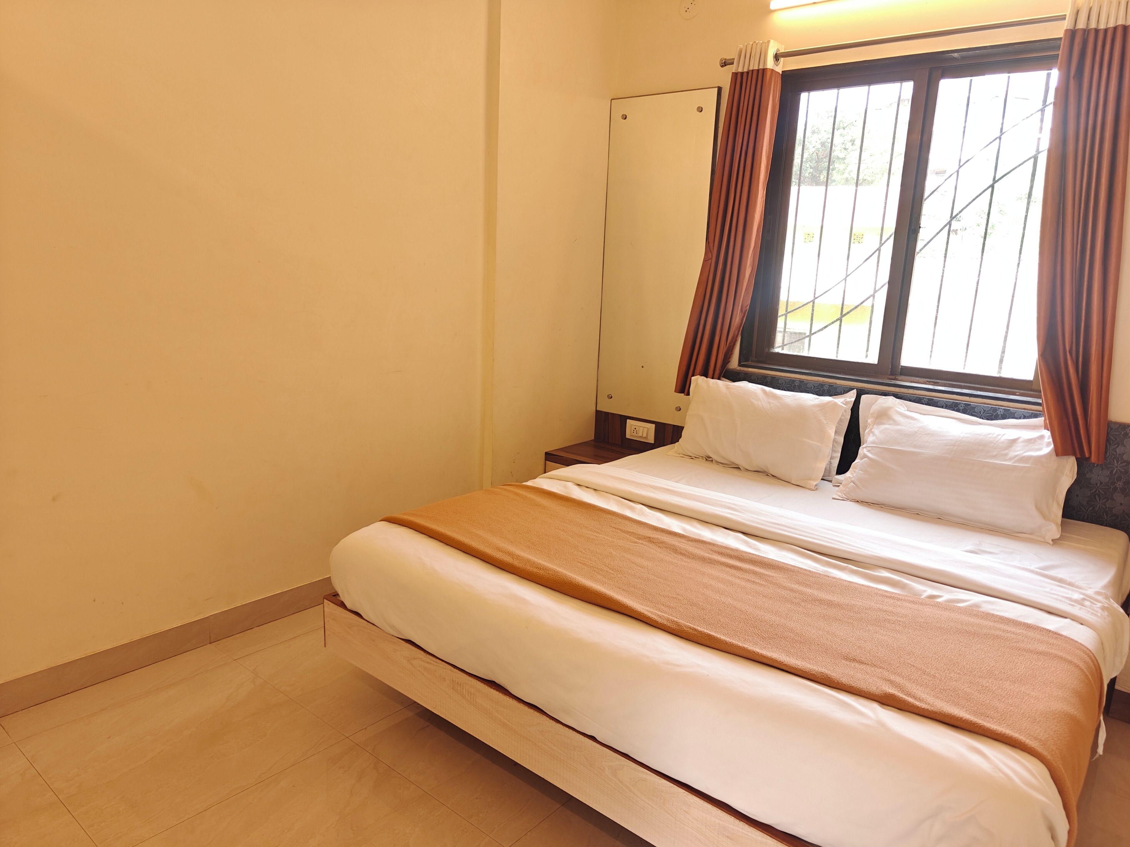 A single bed with a brown blanket, a window with curtains, and a wooden nightstand.