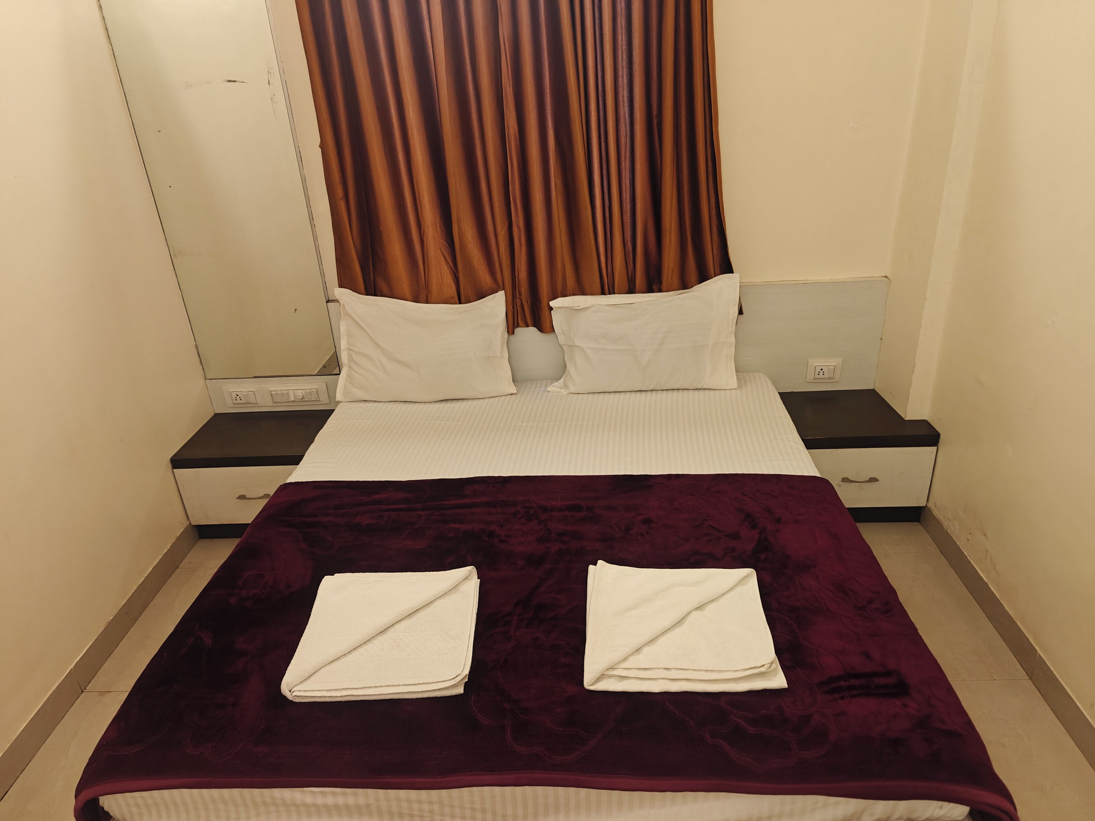 A hotel room with a bed, two pillows, a nightstand, a drawer, and a curtain.