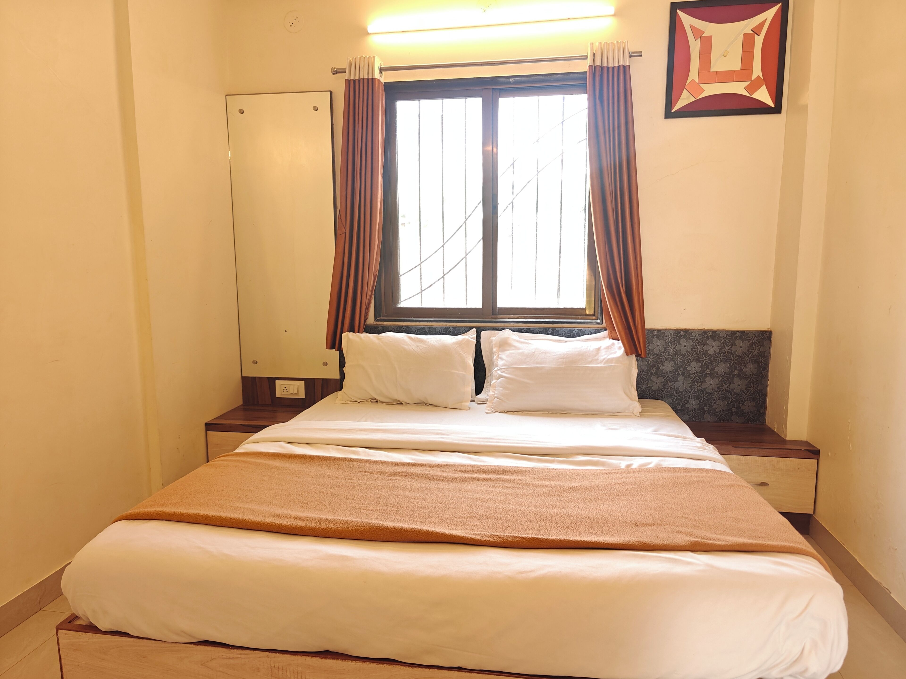 A hotel room with a bed, two bedside tables, a window with curtains, a mirror, and a wall-mounted picture.