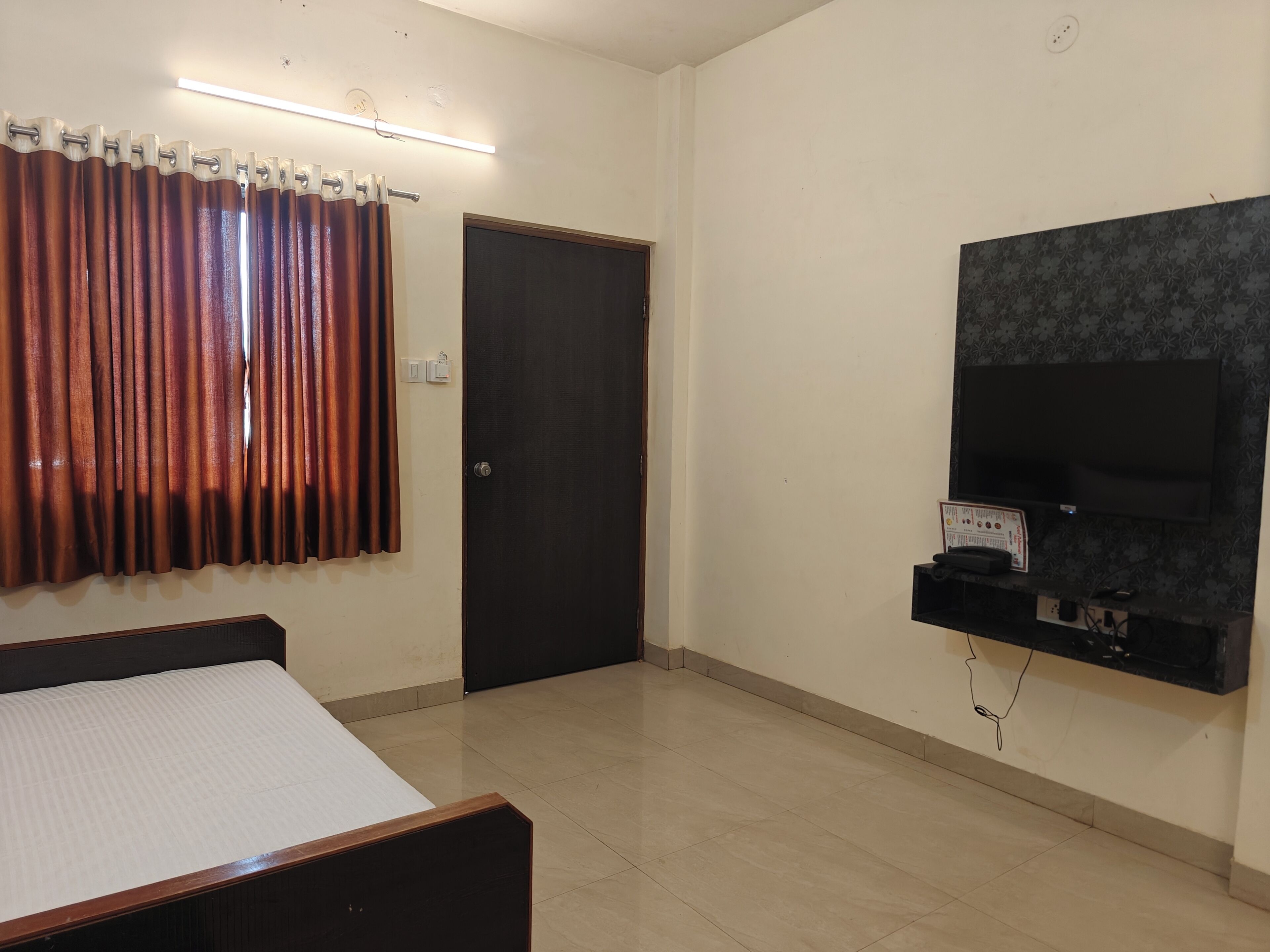 A room with a bed, a dark door, a TV mounted on the wall, and a curtain.
