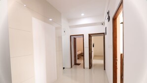 Un pasillo moderno con paredes blancas y suelo de baldosas, que conduce a un dormitorio con puerta de madera y una ventana pequeña.