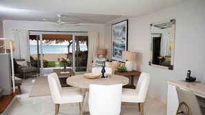A modern hotel room with a dining area, a large window with a view of the beach, a ceiling fan, and a mirror on the wall. - Beachfront Bliss: Ground Floor Condo at The Islands Club, 7 Mile Beach, #05 (Grand Cayman)