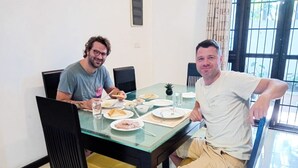 Two people seated at a glass-top dining table with plates of food, chairs, and a window with curtains. - Zara Home (Galle)
