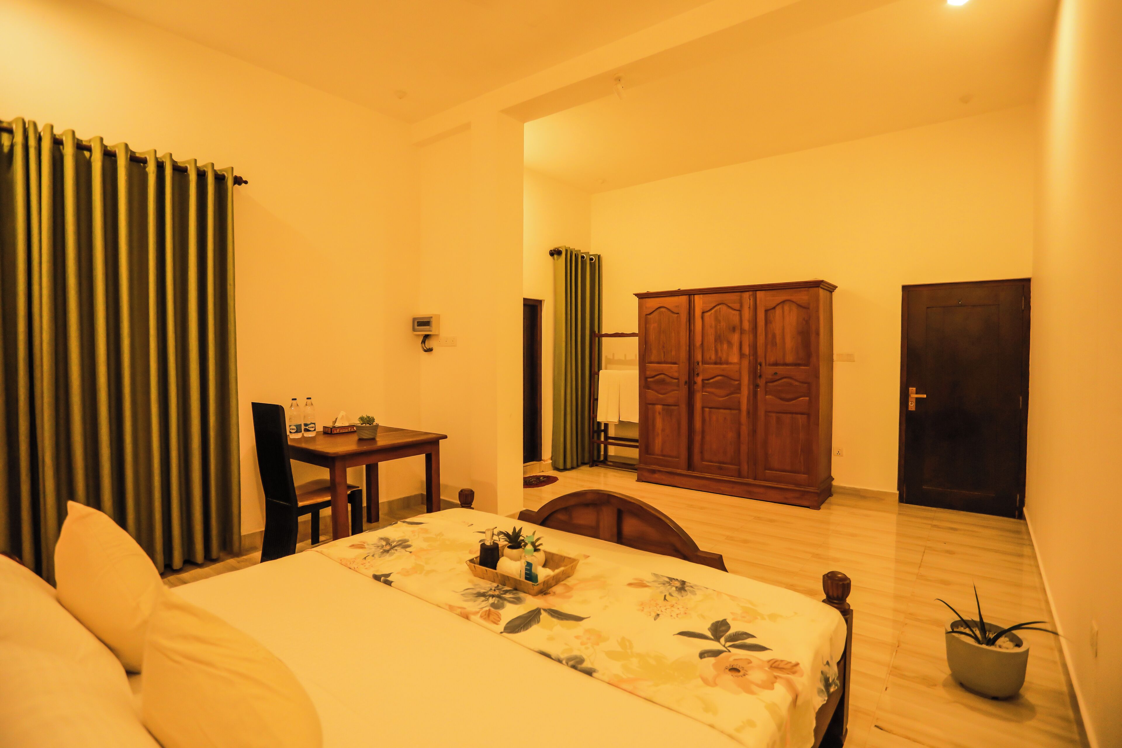 A hotel room with a bed, a wooden desk, a chair, a wardrobe, and a potted plant.