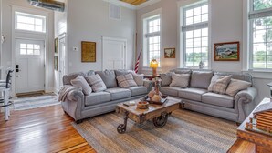 A spacious living room with a large gray sectional sofa, a wooden coffee table, and a view of the outdoors through large windows. - West Lafayette Home ~ 3 Mi to Purdue Campus! (West Lafayette)