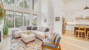 Amplia sala de estar con un sofá modular grande, una mesa de centro de madera y una alfombra estampada. La habitación cuenta con ventanas grandes con vistas a árboles y una cocina moderna con armarios blancos y electrodomésticos de acero inoxidable.