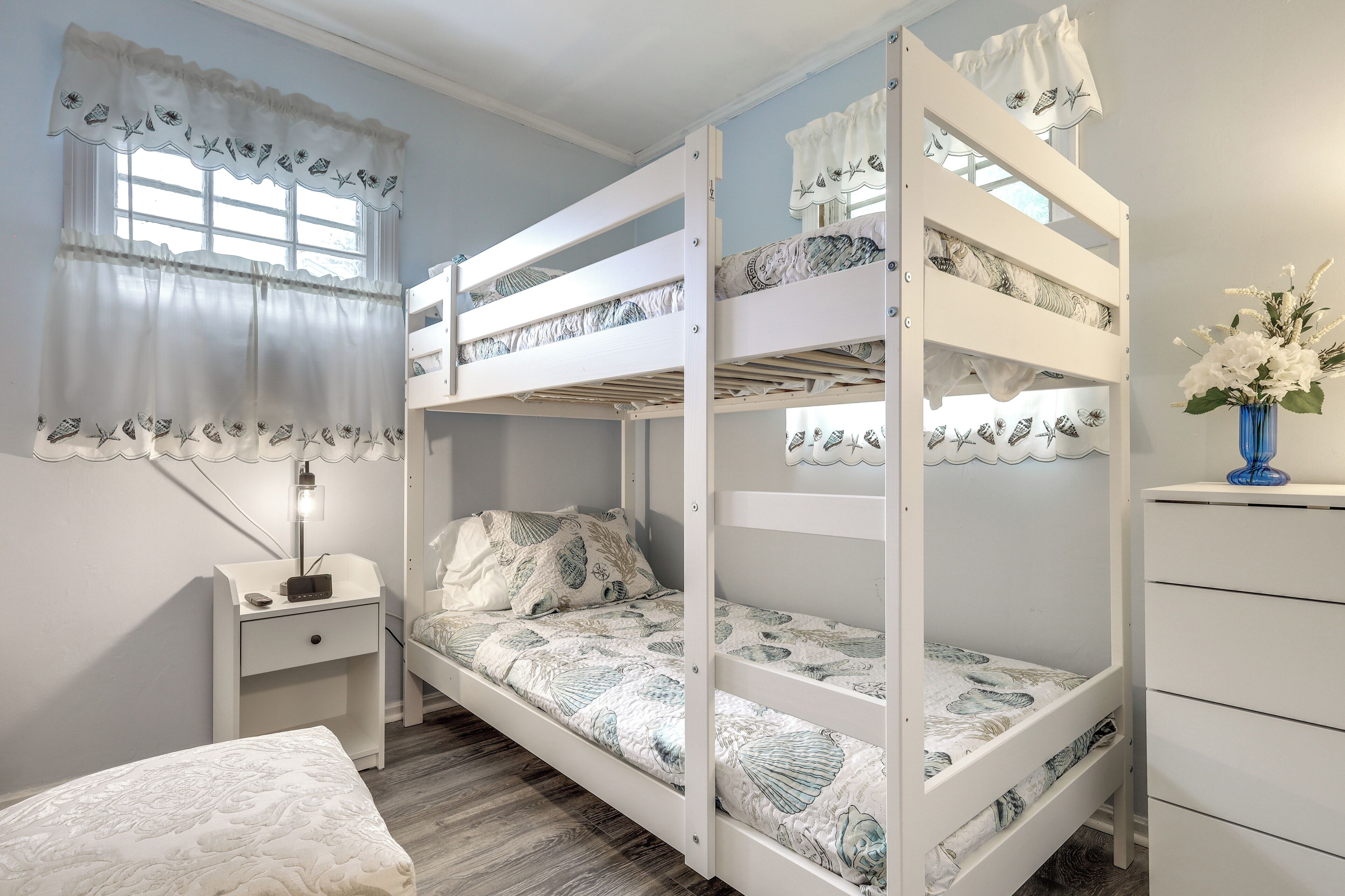 A bunk bed with a white frame and patterned bedding, a bedside table with a lamp, and a dresser with a vase of flowers.