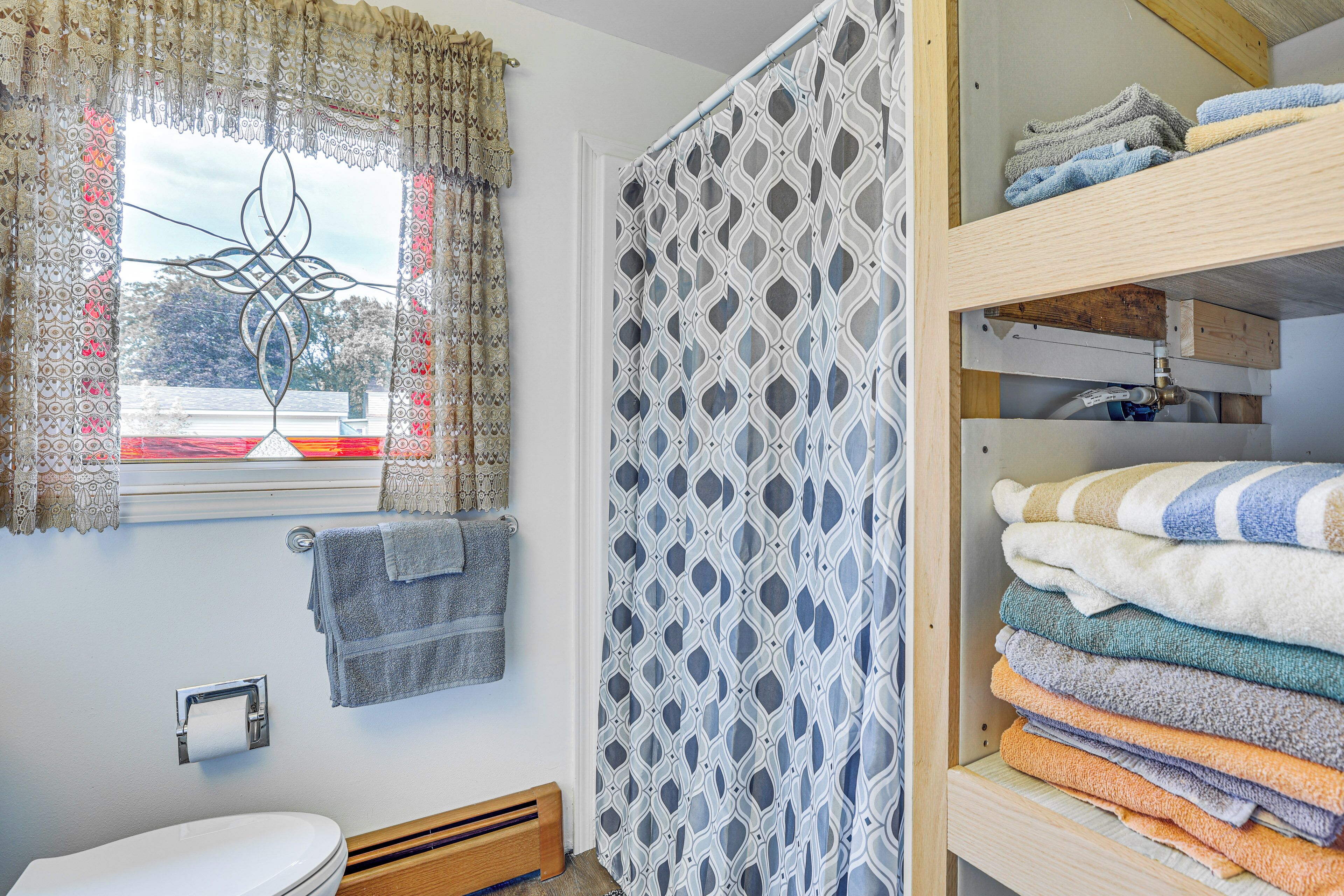 Un baño con un inodoro, un perchero con toallas dobladas, una cortina de ducha estampada y un estante de madera con toallas apiladas.