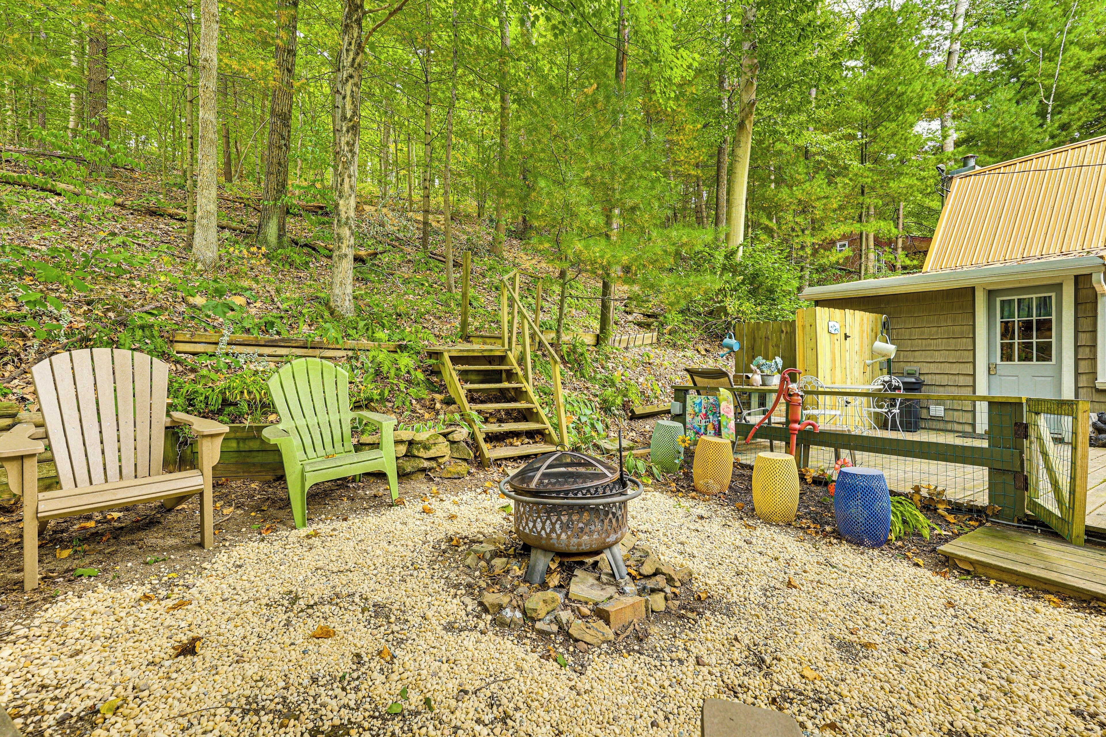 A rustic cabin with a gravel patio, a fire pit, wooden Adirondack chairs, and a wooden staircase leading into the forest.