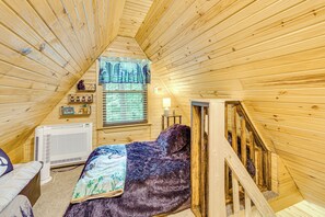 Een gezellig slaapkamer onder het dak, met een houten schuine hemel, een bed met een paarse deken, een raam met zonwering, een verwarming en een klein tafeltje met decoratieve objecten.