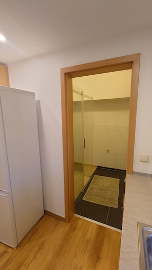 Una pequeña cocina con un refrigerador, una puerta de madera que da a un baño con mampara de vidrio, y una pequeña alfombra en el suelo.