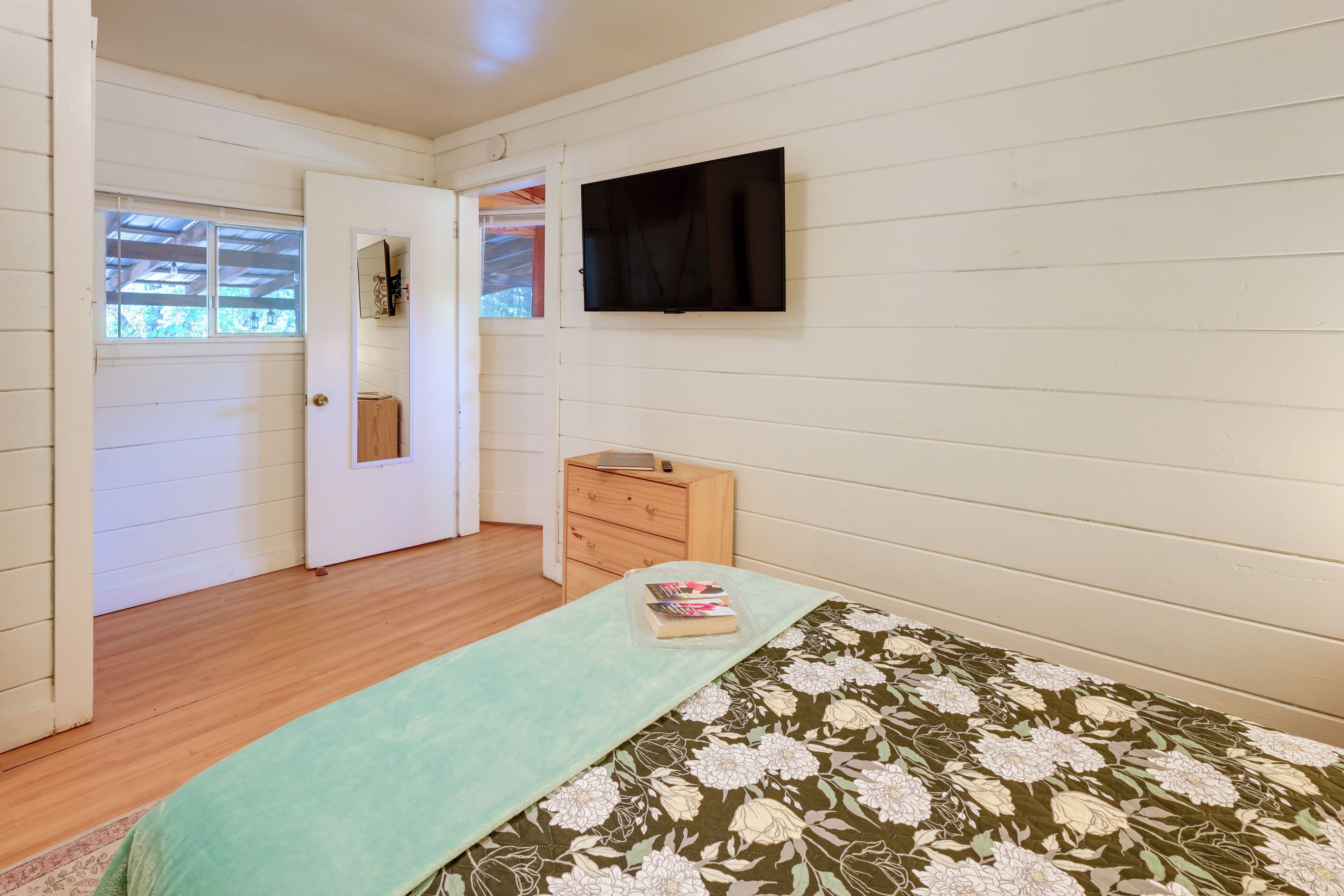A room with a bed, a wooden dresser, a TV mounted on the wall, and a door leading to another room.