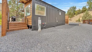 A modern cabin with a large window, wooden exterior, and a gravel driveway.