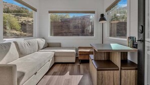 A modern, compact living space with a sofa, a small desk, and a view of a natural landscape through large windows.