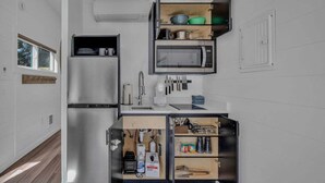 Cocina compacta con microondas integrado, refrigerador y estante con vajilla y utensilios.